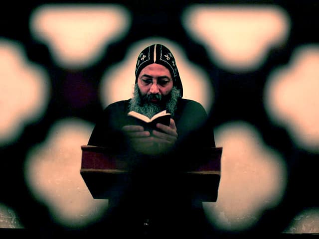Ein bärtiger Priester betet mit der Bibel in der Hand hinter einer ornamental verzierten Mauer. Ein bärtiger Priester betet mit der Bibel in der Hand hinter einer ornamental verzierten Mauer.