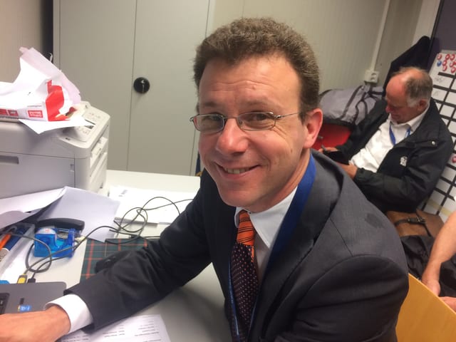 Ein Mann mit Brille sitzt an einem Schreibpult und lacht in die Kamera. Ein Mann mit Brille sitzt an einem Schreibpult und lacht in die Kamera.