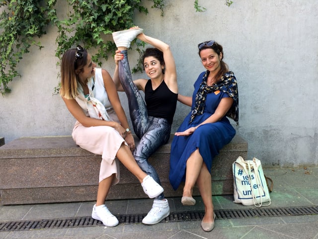 Three women are sitting on a bench, one stretching their leg in the air. Three women are sitting on a bench, one stretching their leg in the air.