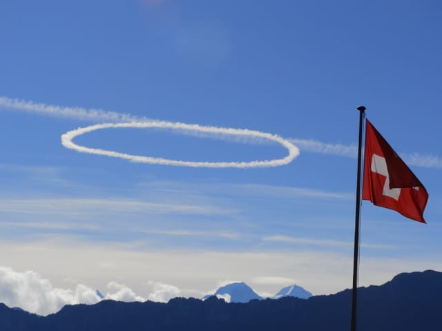 Eine kreisrunde Wolke am sonst blauen Himmel. Im Vordergrund weht die Schweizer Flagge im Wind.