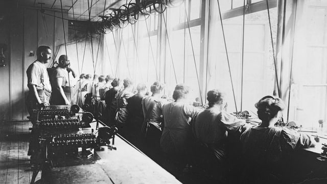 Einblick in eine Uhrenfabrik. Arbeiter sitzen an einer langen Werkbank. Einblick in eine Uhrenfabrik. Arbeiter sitzen an einer langen Werkbank.