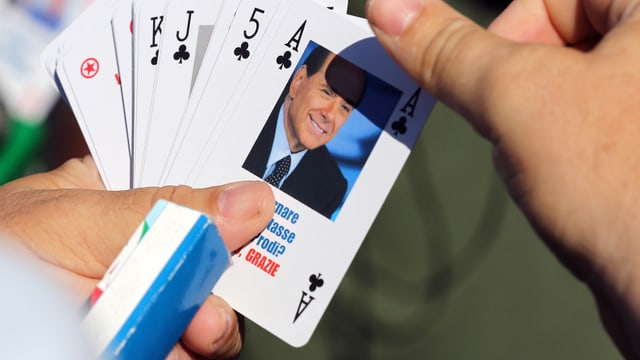 Eine Hand hält Spielkarten in der Hand. Eine davon trägt das Konterfei Silvio Belrusconis. Eine Hand hält Spielkarten in der Hand. Eine davon trägt das Konterfei Silvio Belrusconis.