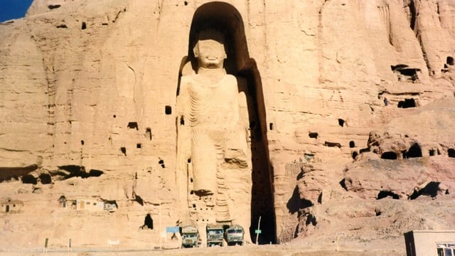 Buddhastatue in einer Felsnische. Davor stehen Autos. Sie sehen munzig aus. Buddhastatue in einer Felsnische. Davor stehen Autos. Sie sehen munzig aus.