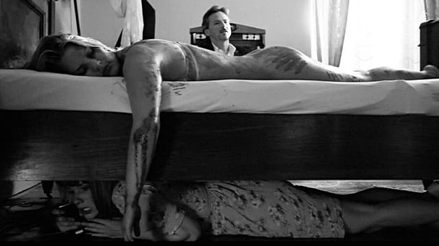 Eine blutige Frau liegt auf dem Bett. Unter dem Bett versteckt sich eine andere Frau. Und im Hintergrund sitzt ein Mann im Anzug. Eine blutige Frau liegt auf dem Bett. Unter dem Bett versteckt sich eine andere Frau. Und im Hintergrund sitzt ein Mann im Anzug.