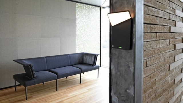 Blick in ein Zimmer mit Holzboden und einem Sofa. Im Vordergrund ist eine HAuswand aus brauenem, gepresstem MAterial zu sehen,