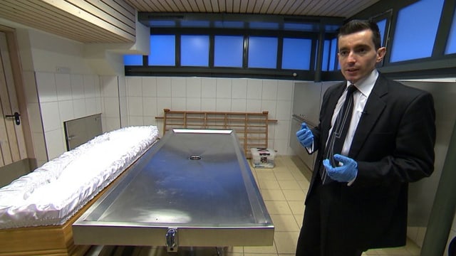 Enver Fazliji trägt einen Anzug und blaue Handschuhe, er steht in einem Leichenaufbahrungsraum. Enver Fazliji trägt einen Anzug und blaue Handschuhe, er steht in einem Leichenaufbahrungsraum.