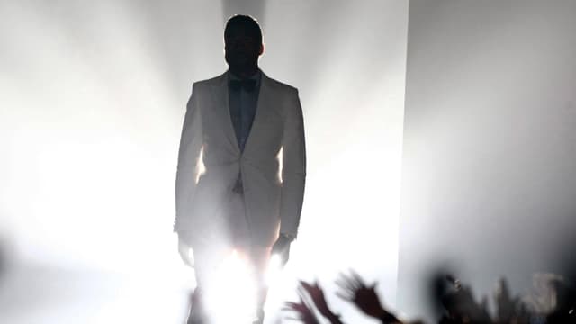 Ein Mann in weissem Anzug schreitet durch grelles Licht, unter ihm versuchen Hände nach ihm zu greifen. Ein Mann in weissem Anzug schreitet durch grelles Licht, unter ihm versuchen Hände nach ihm zu greifen.
