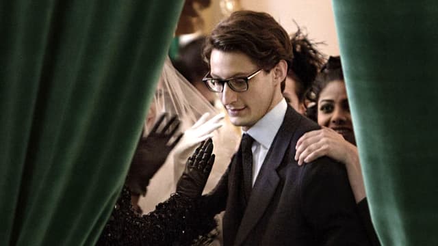 Durch eine Öffnung eines grünen Vorhangs sieht man einen jungen Mann, der von Frauen beklatscht wird. Durch eine Öffnung eines grünen Vorhangs sieht man einen jungen Mann, der von Frauen beklatscht wird.