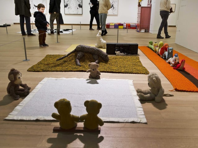 Eine Installation des des US-amerikanischen Künstler Mike Kelley im Stedelijk Museum in Amsterdam, 2012: Stoffbären sitzen sich in einem Quadrat gegenüber, dahinter sind zwei Leoparden und weitere Tiere in einer Reihe zu sehen.