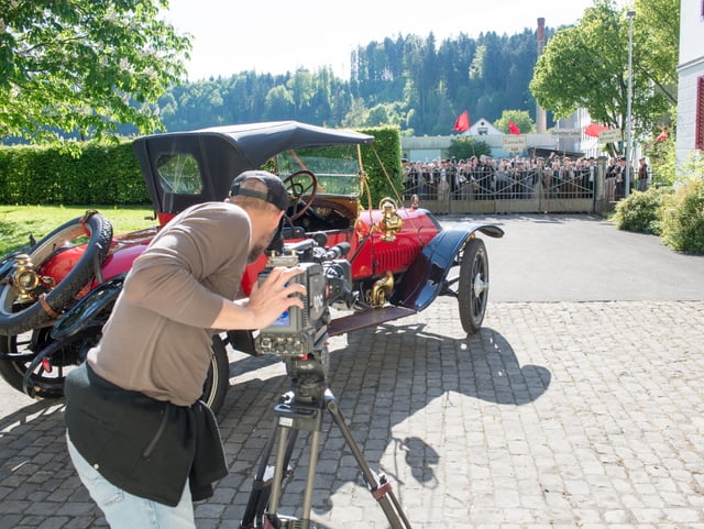 Kamermann filmt im Vordergrund das neue Auto, im Hintergrund die Arbeiterdemonstration