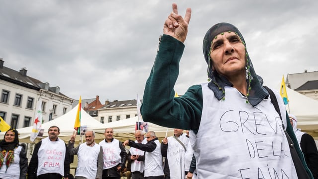 Im Vordergrund eine ältere Frau, sie macht ein Victory-Zeichen und trägt ein Shirt, auf dem steht «grève de faim», hinter ihr weitere Frauen und Männer mit gleichen Shirts. Im Vordergrund eine ältere Frau, sie macht ein Victory-Zeichen und trägt ein Shirt, auf dem steht «grève de faim», hinter ihr weitere Frauen und Männer mit gleichen Shirts.
