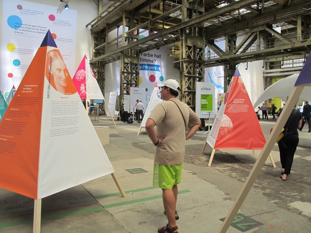 Blick in die Halle in der mit Holzstangen Stoffpyramiden mit aufgedruckten Informationen aufgestellt sind. Blick in die Halle in der mit Holzstangen Stoffpyramiden mit aufgedruckten Informationen aufgestellt sind.