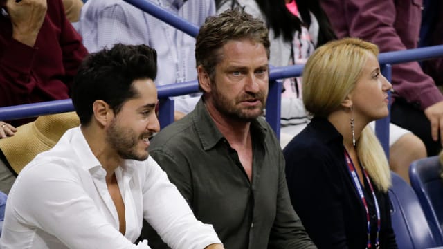 Gerard Butler sitzt auf den Zuschauerrängen und schaut angespannt aufs Feld, links ein Mann, rechts eine Frau.