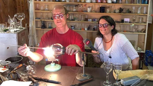 Ein Mann bearbeitet über einer Flamme Glas, daneben sitzt eine Frau mit SRF-Mikrofon. Beide tragen eine Schutzbrille. Ein Mann bearbeitet über einer Flamme Glas, daneben sitzt eine Frau mit SRF-Mikrofon. Beide tragen eine Schutzbrille.