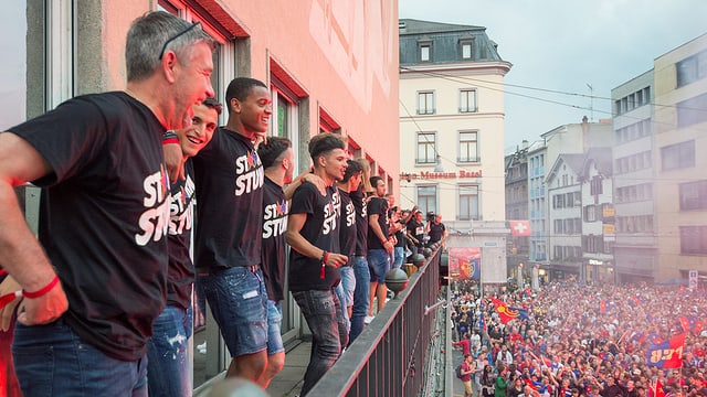 Männer stehen auf Balkon, zahlreiche Menschen feiern auf dem darunterliegenden Platz Männer stehen auf Balkon, zahlreiche Menschen feiern auf dem darunterliegenden Platz