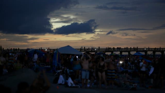 Viele Menschen stehen an einem Stand in den USA mit Blick zur abgedunkelten Sonne. Viele Menschen stehen an einem Stand in den USA mit Blick zur abgedunkelten Sonne.