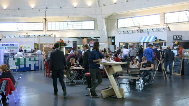 Blick in den Innenraum der Basler Markthalle mit Essensständen, Tischen und Stühlen. Viele Leute sind am Essen. Blick in den Innenraum der Basler Markthalle mit Essensständen, Tischen und Stühlen. Viele Leute sind am Essen.