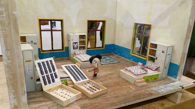 Eine Puppe in einer Art Puppenhaus, davor Boxen mit Puppen-Elementen Eine Puppe in einer Art Puppenhaus, davor Boxen mit Puppen-Elementen