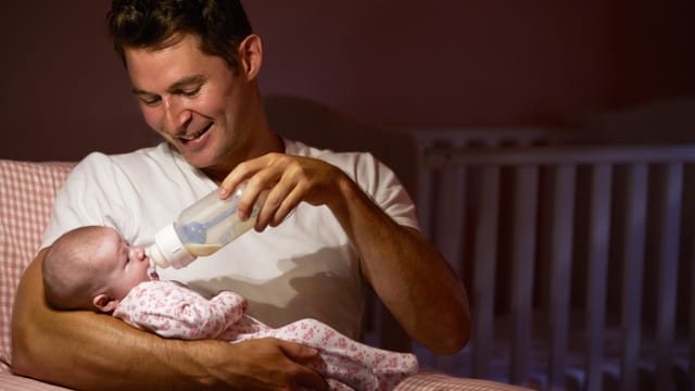 Ein Mann sitzt lächelnd auf einem Stuhl und gibt seinem Baby, das er auf dem Arm hält, ein Fläschen. Ein Mann sitzt lächelnd auf einem Stuhl und gibt seinem Baby, das er auf dem Arm hält, ein Fläschen.