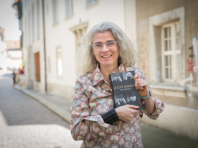 Eine Frau mit Brille steht in einer Altstadtgasse und hält ein dickes Buch.