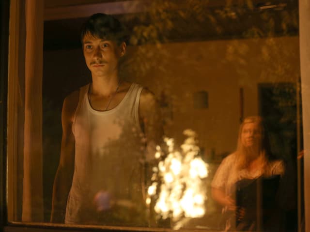Ein Feuer brennt. Ein Mann steht am Fenster, eine Frau steht auf draussen. Sie blicken sich gegenseitig an. Ein Feuer brennt. Ein Mann steht am Fenster, eine Frau steht auf draussen. Sie blicken sich gegenseitig an.