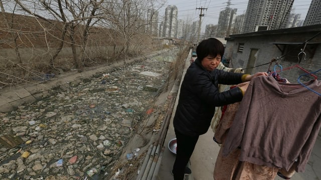 Frau hängt die Wäsche auf, hinter ihr fliesst ein Fluss durch, an dessen Oberfläche lauter Plastik schwimmt Frau hängt die Wäsche auf, hinter ihr fliesst ein Fluss durch, an dessen Oberfläche lauter Plastik schwimmt