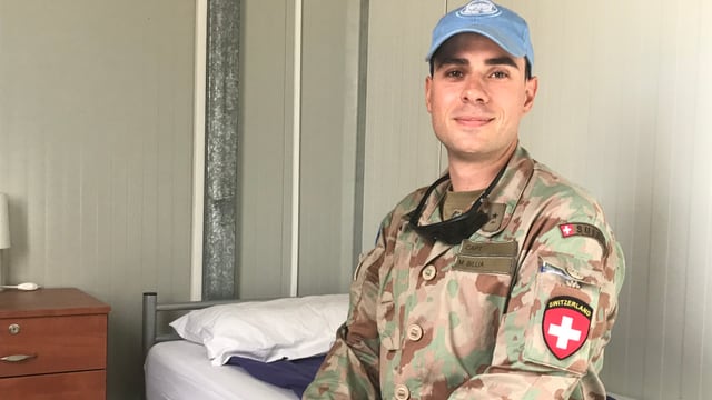 Mann mit blauer Dächlikappe im Kampfanzug und Abzeichen. Er sitzt auf einem Bett. Mann mit blauer Dächlikappe im Kampfanzug und Abzeichen. Er sitzt auf einem Bett.