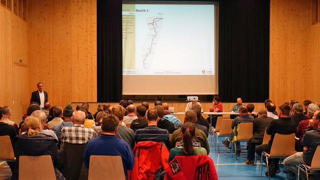 Ein Saal, einige Dutzend Personen sitzen auf Stühlen, vorne steht ein Mann und spricht, auf einer Leinwand ist eine Strassenkarte sichtbar. Ein Saal, einige Dutzend Personen sitzen auf Stühlen, vorne steht ein Mann und spricht, auf einer Leinwand ist eine Strassenkarte sichtbar.
