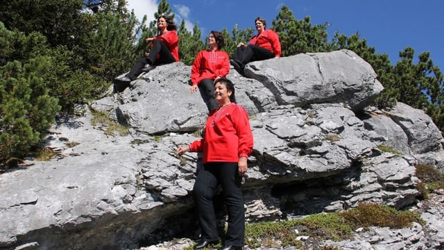 Die Sängerinnen tragen alle rote Blusen und sitzen auf einem grossen Stein inmitten der Bergwelt. Die Sängerinnen tragen alle rote Blusen und sitzen auf einem grossen Stein inmitten der Bergwelt.