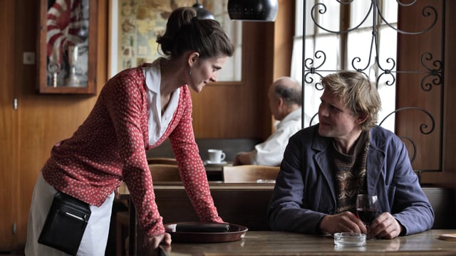 Filmszene: Ernst sitzt in einer Beiz am Tisch, die Kellnerin Regula stützt sich auf den Tisch und spricht mit Ernst.