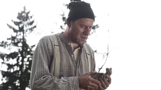 Ein Mann mit Hemd, Hosenträgern und Zipfelmütze schaut auf eine verfaulte Kartoffel in seiner Hand. Im Hintergrund sieht man ein paar Tannen. Ein Mann mit Hemd, Hosenträgern und Zipfelmütze schaut auf eine verfaulte Kartoffel in seiner Hand. Im Hintergrund sieht man ein paar Tannen.