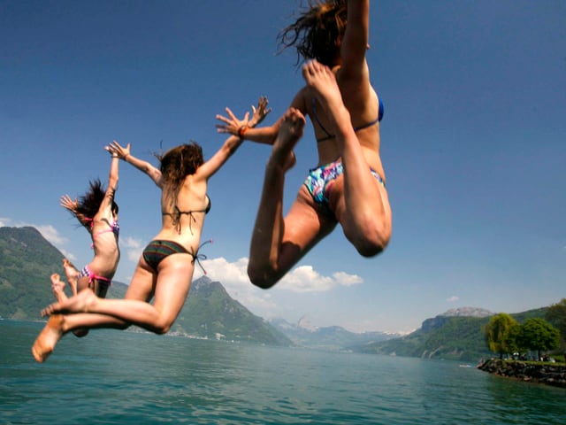 See bei sonnigem Wetter, im Hintergrund Ufer mit Bergen. Im Vordergrund drei Mädchen im Bikini, die ins Wasser springen. See bei sonnigem Wetter, im Hintergrund Ufer mit Bergen. Im Vordergrund drei Mädchen im Bikini, die ins Wasser springen.