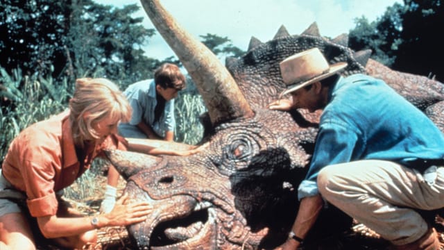 Ein Dinosaurier liegt am Boden, ein Mann, eine Frau und ein Junge stehen um ihn herum und streicheln ihm den Kopf. Ein Dinosaurier liegt am Boden, ein Mann, eine Frau und ein Junge stehen um ihn herum und streicheln ihm den Kopf.