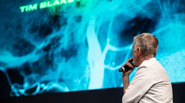 Cooper steht vor einer Leinwand, auf der ein nicht genau erkennbares, blaues Bild zu sehen ist. Cooper steht vor einer Leinwand, auf der ein nicht genau erkennbares, blaues Bild zu sehen ist.