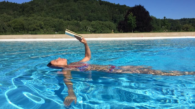 Annette König schwimmt in einem Pool auf dem Rücken und hält vor sich ausgestreckt über dem Wasser ein gelbes Buch in ihren Händen