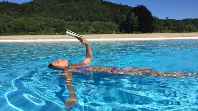Annette König schwimmt in einem Pool auf dem Rücken und hält vor sich ausgestreckt über dem Wasser ein gelbes Buch in ihren Händen Annette König schwimmt in einem Pool auf dem Rücken und hält vor sich ausgestreckt über dem Wasser ein gelbes Buch in ihren Händen