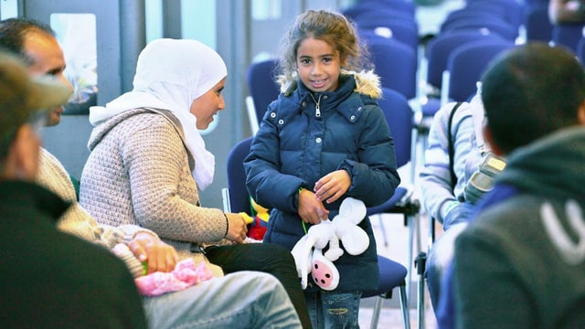 Ein Mäschen mit einer Diddlemaus in der rechten Hand lächelt, links von ihr eine Frau mit Kopftuch auf einem Stuhl. Ein Mäschen mit einer Diddlemaus in der rechten Hand lächelt, links von ihr eine Frau mit Kopftuch auf einem Stuhl.