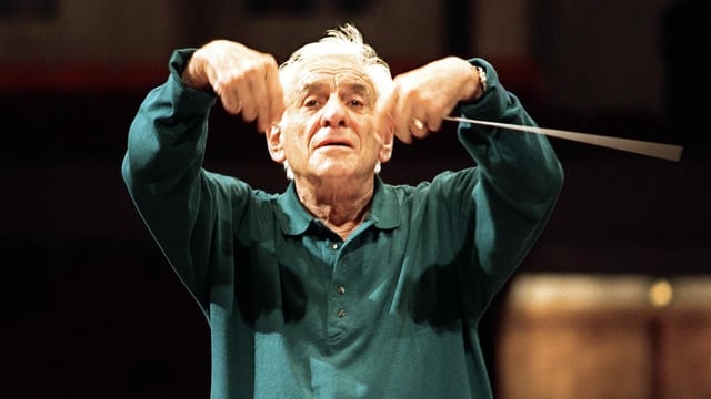 Älterer Mann mit grauen Haaren und dunkelgrünem Shirt dirigiert Orchester während einer Probe Älterer Mann mit grauen Haaren und dunkelgrünem Shirt dirigiert Orchester während einer Probe