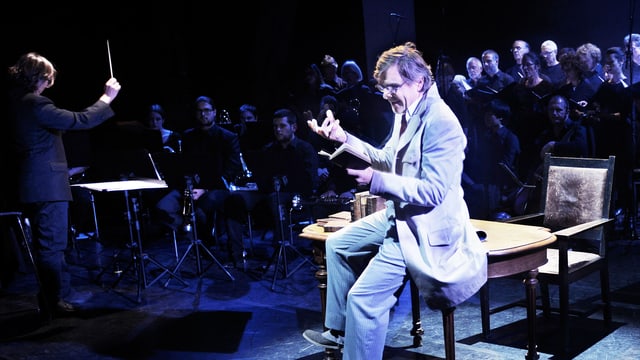 Ein Mann sitzt auf einem Stuhl. Hinter ihm ist ein Orchester. Ein Mann sitzt auf einem Stuhl. Hinter ihm ist ein Orchester.