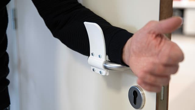 An einer Türfalle ist ein Plastikverlängerung angebracht, an der jemand mit dem Arm zieht. An einer Türfalle ist ein Plastikverlängerung angebracht, an der jemand mit dem Arm zieht.