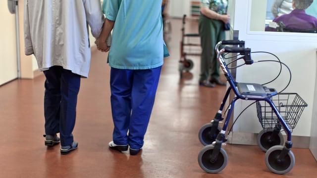Pflegerin begleitet älteren Menschen durch einen Gang, im Vordergrund ein abgestellter Rollator. Pflegerin begleitet älteren Menschen durch einen Gang, im Vordergrund ein abgestellter Rollator.