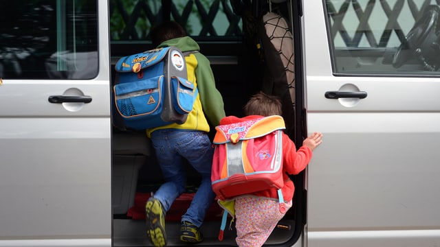 Zwei Kinder steigen in ein grosses graues Auto ein. Sie kommen von der Schule. Das sieht man an den Schul-Rucksäcken. Zwei Kinder steigen in ein grosses graues Auto ein. Sie kommen von der Schule. Das sieht man an den Schul-Rucksäcken.