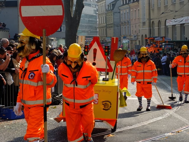 Männer in orangen Overalls mit Verkehrsschildern und Besen.