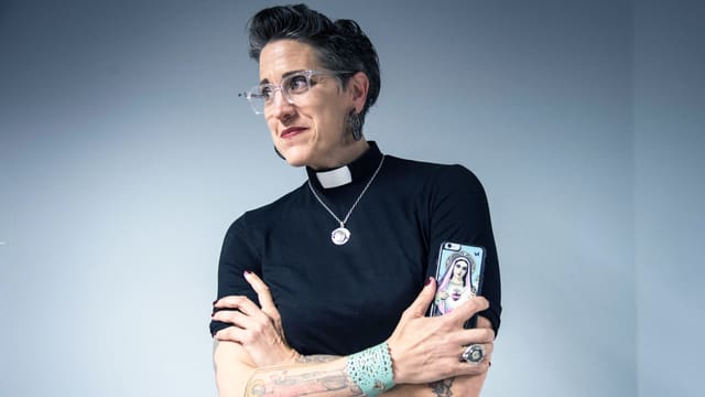 Eine Frau mit Kurzhaarschnitt und Tattoos hält ein Smartphone in der Hand. Sie ist Pfarrerin und trägt ein schwarzes T-Shirt, am Hals ist eine weisse Pfarrersbinde und eine Goldkette. Eine Frau mit Kurzhaarschnitt und Tattoos hält ein Smartphone in der Hand. Sie ist Pfarrerin und trägt ein schwarzes T-Shirt, am Hals ist eine weisse Pfarrersbinde und eine Goldkette.