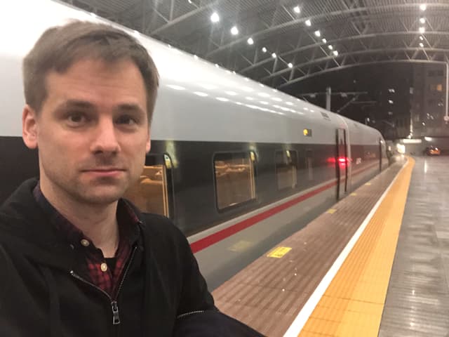 China-Korrespondent Martin Aldrovandi macht ein Selfie am Perron eines Chinesischen Bahnhofes. Hinter ihm steht ein grauer chinesischer Schnellzug mit roten Streifen auf der Seite. China-Korrespondent Martin Aldrovandi macht ein Selfie am Perron eines Chinesischen Bahnhofes. Hinter ihm steht ein grauer chinesischer Schnellzug mit roten Streifen auf der Seite.