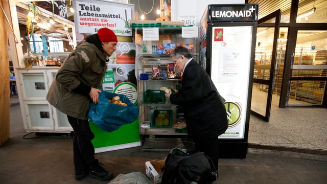 Ein junger Mann und eine ältere Frau stehen vor einem offenen Kühlschrank, der vor einem Supermarkt platziert ist. Ein junger Mann und eine ältere Frau stehen vor einem offenen Kühlschrank, der vor einem Supermarkt platziert ist.