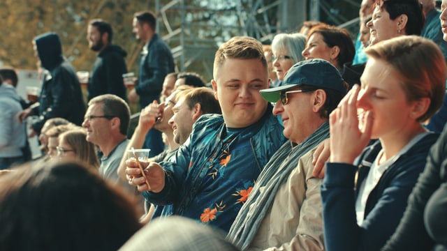 Im Fokus: Zwei Männer sitzend auf einer Fussballtribüne. Einer ist jung, der andere älter. Der ältere Mann trägt Mütze und Sonnenbrille. Beide lächeln. Im Fokus: Zwei Männer sitzend auf einer Fussballtribüne. Einer ist jung, der andere älter. Der ältere Mann trägt Mütze und Sonnenbrille. Beide lächeln.