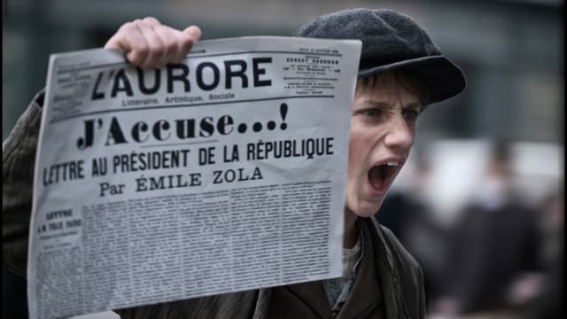 Zeitungsjunge hält schreiend eine Zeitunng in die Luft, auf der der Titel «J'accuse!» zu lesen ist. Zeitungsjunge hält schreiend eine Zeitunng in die Luft, auf der der Titel «J'accuse!» zu lesen ist.