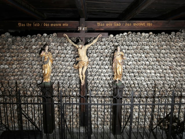 Figur von Jesus am Kreuz, dahinter Wand mit Totenschädeln Figur von Jesus am Kreuz, dahinter Wand mit Totenschädeln