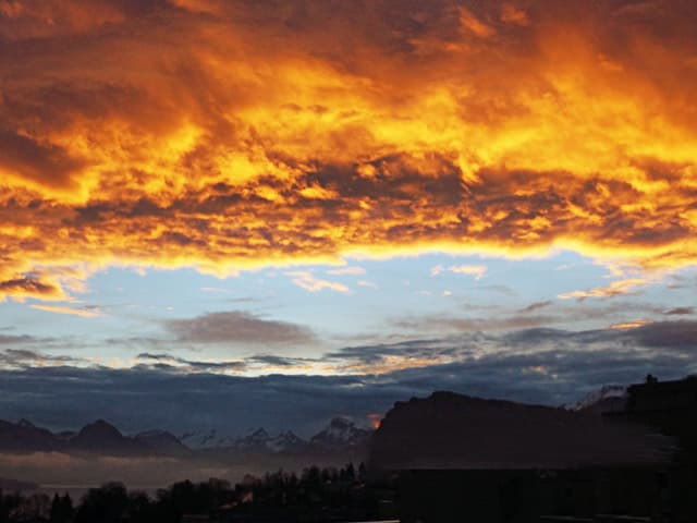 Die Wolken am Himmel leuchten orange und rot. Darunter liegen die Bergen noch im Halbdunkeln.